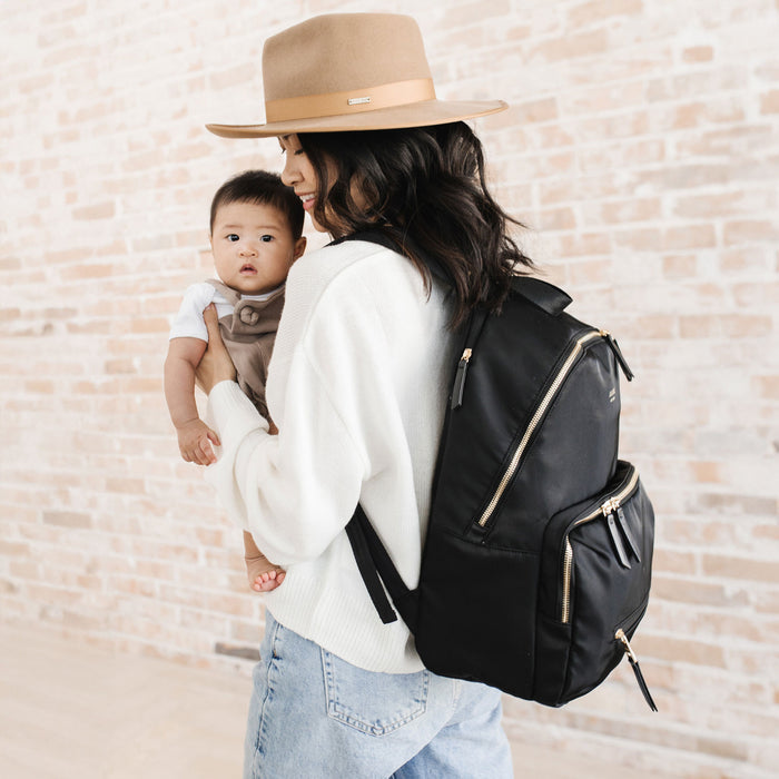 Woman holds baby while wearing JuJuBe Everyday Backpack - Black