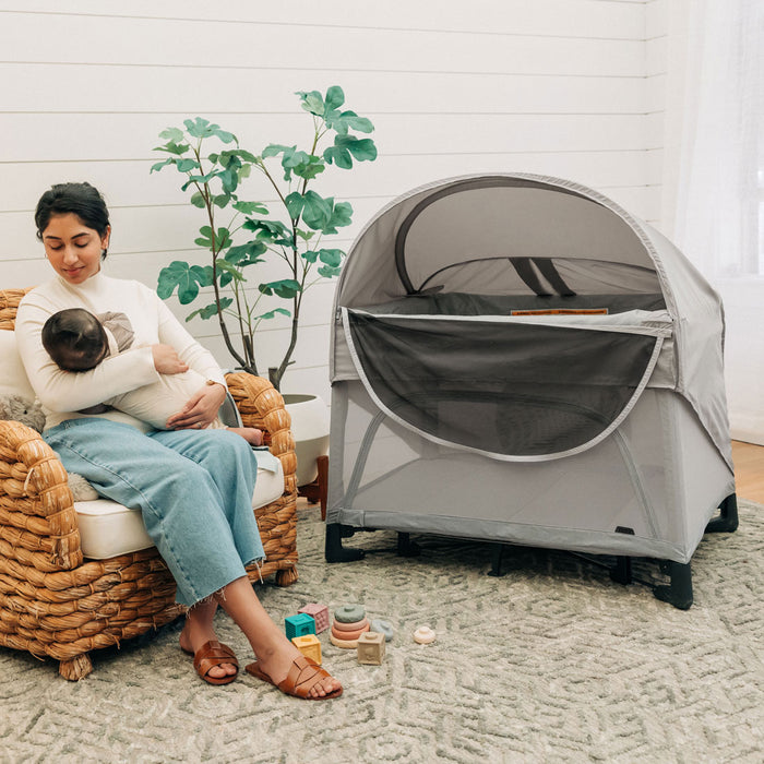 Mom holding baby sits next to REMI playard with attached UPPAbaby REMI Playard Canopy